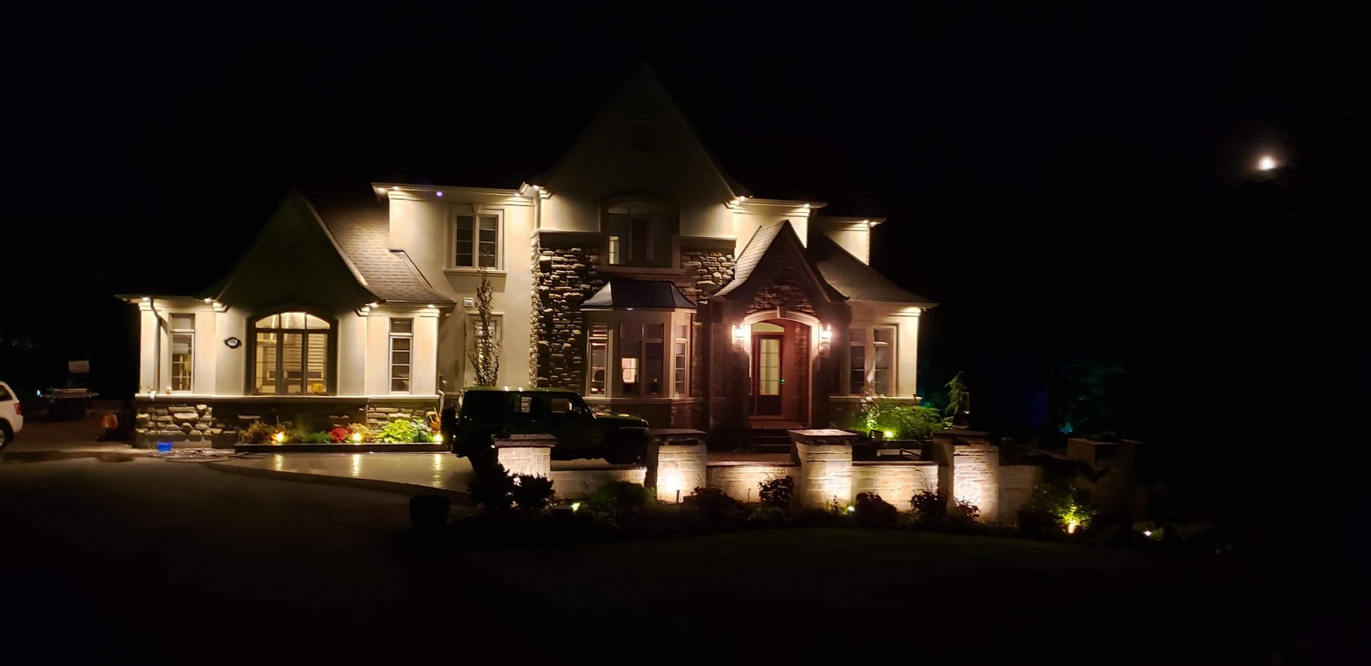 Two-storey home exterior at night lit by soffit lighting and illuminated stone garden walls